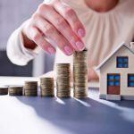 A person stacking coins beside a miniature house, symbolizing real estate investment and housing market trends.
